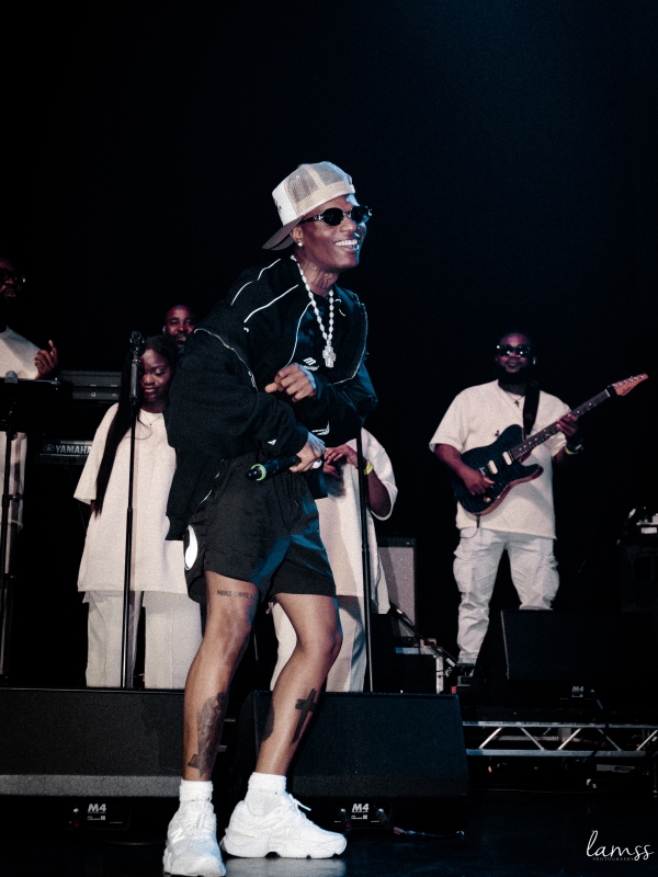 Wizkid in a black jacket and shorts dancing on stage with a live band and backing vocalists in the background. — concert photography London by Lamss Media